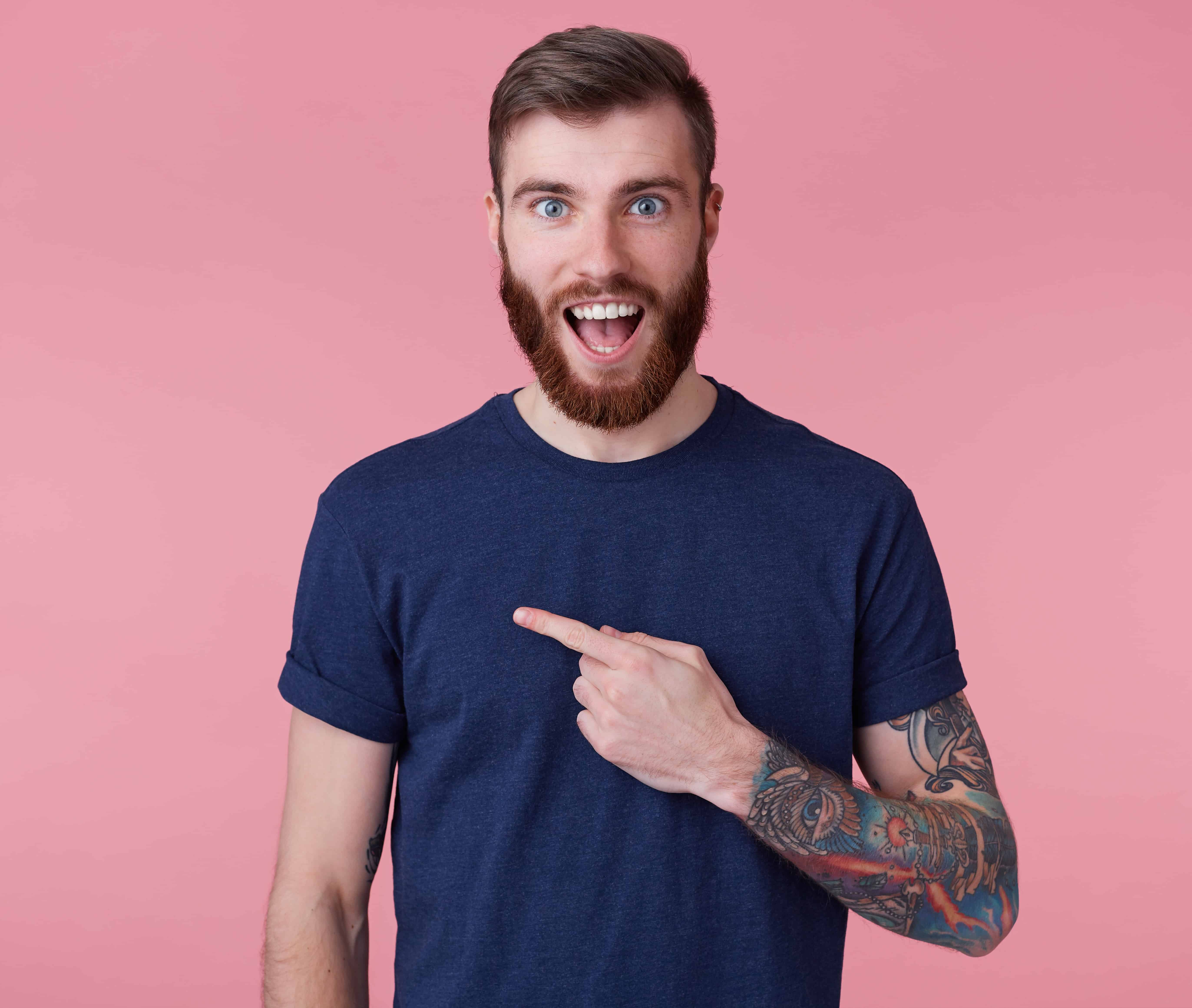 Young happy amazed attractive red-bearded young guy , wearing a blue t-shirt, with wide open mouth in surprise, pointing finger to copy space at the left side isolated over pink background. Young happy amazed attractive red-bearded young guy , wearing a blue t-shirt, with wide open mouth in surprise, pointing finger to copy space at the left side isolated over pink background.